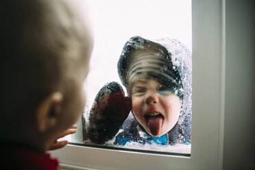 Seitenansicht eines kleinen Jungen, der seinen Bruder betrachtet, der im Winter durch das Fenster seine Zunge herausstreckt - CAVF56617