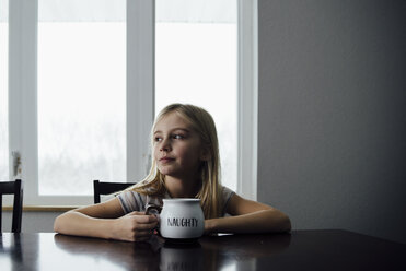 Nachdenkliches Mädchen, das eine Tasse hält, während es auf einem Stuhl am Tisch zu Hause sitzt - CAVF56618