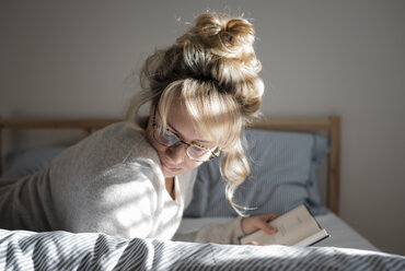 Seitenansicht einer Frau, die ein Buch liest, während sie zu Hause auf dem Bett liegt - CAVF56648