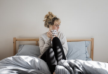 Frau hält Kaffeetasse, während sie zu Hause auf dem Bett sitzt - CAVF56650