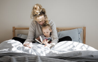 Mother with daughter reading book while sitting on bed at home - CAVF56651