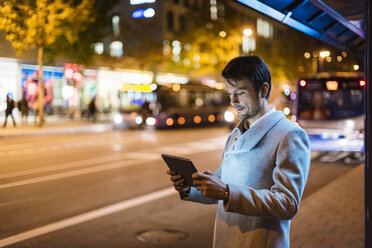 Businessman with digital tablet standing at a bus stop at night - DIGF05559