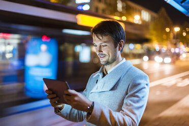 Businessman with digital tablet standing at a bus stop at night - DIGF05561