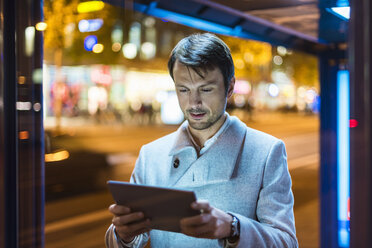 Businessman with digital tablet standing at a bus stop at night - DIGF05562