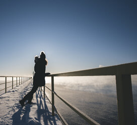 Frau in voller Länge auf einem schneebedeckten Steg mit Geländer über einem zugefrorenen See gegen den Himmel stehend - CAVF56684