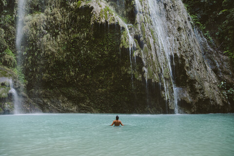 Rückansicht eines Mannes ohne Hemd, der im Fluss bei einem Wasserfall im Wald schwimmt, lizenzfreies Stockfoto