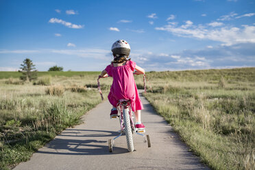 Rear view of girl sitting on bicycle with training wheels against sky during sunny day - CAVF56703