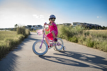 Mädchen fährt Fahrrad mit Stützrädern auf der Straße gegen den Himmel an einem sonnigen Tag - CAVF56704