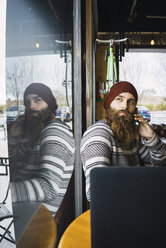 Hipster man answering smart phone while looking through window with laptop computer on table at cafe - CAVF56715