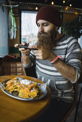 Hipster-Mann, der mit seinem Smartphone im Café das Essen auf dem Teller fotografiert - CAVF56718