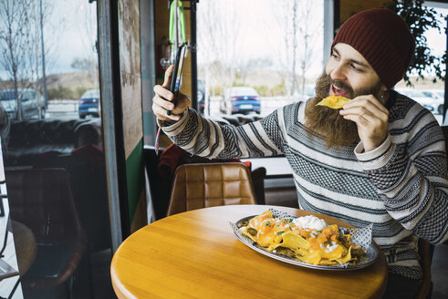 Hipster-Mann, der ein Selfie macht, während er Tortilla-Chips mit seinem Smartphone in einem Café isst - CAVF56719