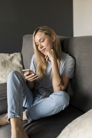 Lächelnde junge Frau sitzt auf der Couch und benutzt ein Mobiltelefon, lizenzfreies Stockfoto