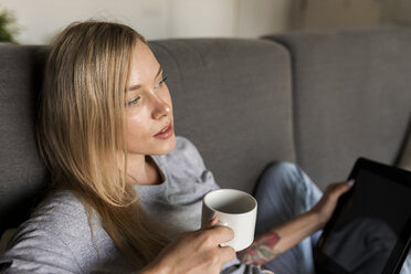 Junge Frau sitzt auf einer Couch und hält eine Kaffeetasse und ein Tablet - VABF01854