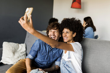 Happy couple sitting on couch taking a selfie - VABF01864