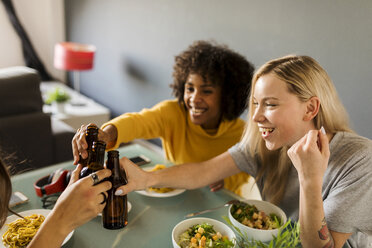 Happy girlfriends sitting at dining table clinking beer bottles - VABF01867