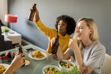 Happy girlfriends sitting at dining table raising beer bottles - VABF01868