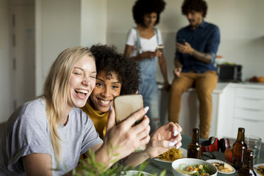 Happy girlfriends with cell phone sitting at dining table - VABF01876