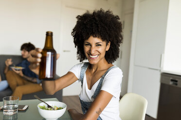 Portrait of smiling woman raising beer bottle at dining table - VABF01892