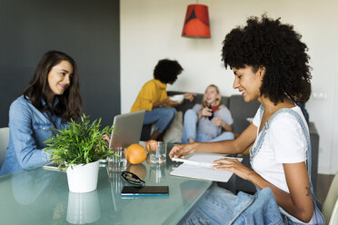 Women using laptop and notebook at dining table with friends in background - VABF01893