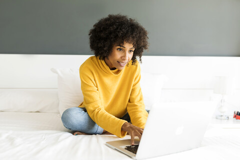 Lächelnde Frau sitzt auf dem Bett und benutzt einen Laptop, lizenzfreies Stockfoto