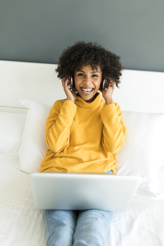 Porträt einer glücklichen Frau, die mit Kopfhörern und Laptop auf dem Bett liegt, lizenzfreies Stockfoto