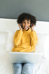 Portrait of happy woman lying on bed with headphones and laptop - VABF01925