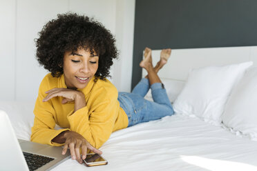 Smiling woman lying on bed using cell phone and laptop - VABF01928