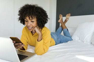 Smiling woman lying on bed using cell phone and laptop - VABF01929