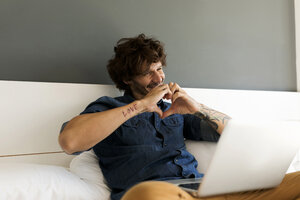 Glücklicher tätowierter Mann mit Laptop, der mit seiner Freundin im Bett plaudert - VABF01936