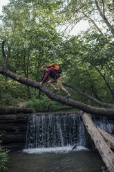 Junger Wanderer mit Rucksack beim Überqueren von Wasser auf einem Baumstamm im Wald - VPIF01177