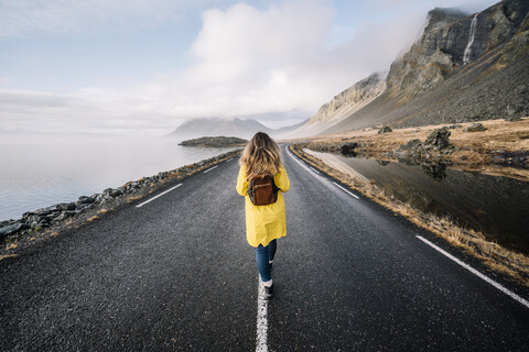 Island, Rückenansicht einer Frau mit Rucksack, die auf dem Mittelstreifen einer Landstraße geht, lizenzfreies Stockfoto