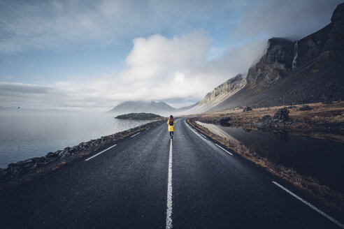 Iceland, back view of woman with backpack walking on median strip of country road - OCMF00121