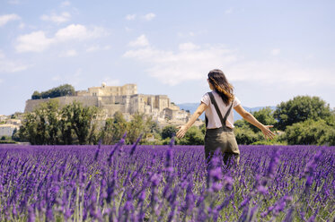 France, Grignan, back view of woman standing in lavender field looking at village - GEMF02594