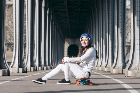 Porträt einer Frau, die auf einem Skateboard auf der Straße unter einer Brücke sitzt, lizenzfreies Stockfoto
