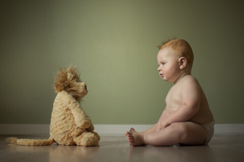 Hemdloser Babyjunge betrachtet Stofftier, während er zu Hause auf dem Boden an der Wand sitzt, lizenzfreies Stockfoto