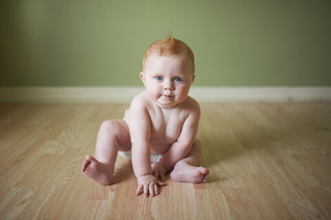 Porträt eines niedlichen, hemdlosen kleinen Jungen, der zu Hause auf dem Boden an der Wand sitzt, lizenzfreies Stockfoto