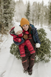 Porträt einer lächelnden Mutter, die ihre Tochter beim Ziehen einer Kiefer im Winter im Wald trägt - CAVF56853