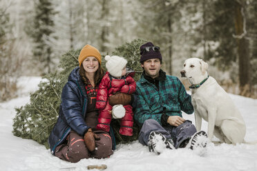 Porträt von Eltern mit Tochter und Hund, die an einer Kiefer auf einem verschneiten Feld im Wald sitzen - CAVF56854