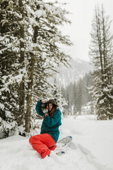 Woman photographing with camera while crouching on snow covered field in forest - CAVF56857