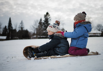 Side view of siblings on sled against cloudy sky - CAVF56876