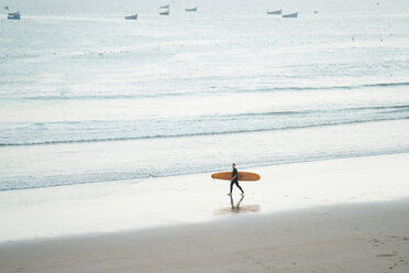 Mann in voller Länge, der ein Surfbrett trägt, während er am Strand spazieren geht - CAVF56934