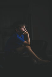 Side view of thoughtful boy with hand on chin sitting on chair in darkroom - CAVF56938