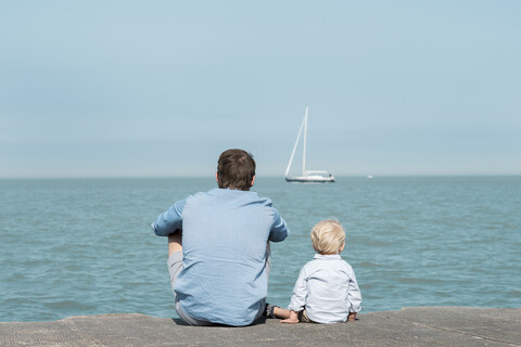 Rückansicht von Vater mit Sohn am Strand sitzend gegen blauen Himmel an einem sonnigen Tag, lizenzfreies Stockfoto