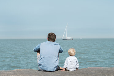 Rear view of father with son sitting at beach against blue sky during sunny day - CAVF56949