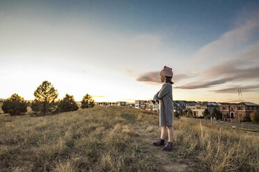 Side view of girl standing on grassy field against sky and cityscape during sunset - CAVF56993