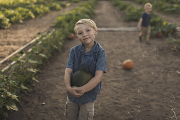 Hochformatiges Porträt eines Jungen, der einen Kürbis trägt, mit seinem Bruder im Hintergrund auf einem Bauernhof - CAVF56996