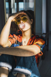 Young woman with head in hand blowing bubble gum at home - CAVF57000