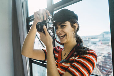 Fröhliche junge Frau beim Fotografieren mit der Kamera, während sie zu Hause am Fenster steht - CAVF57003