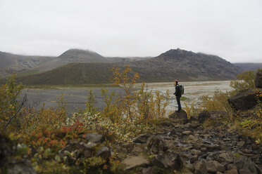 Wanderer in voller Länge mit Blick auf die Aussicht, während er auf einem Felsen am Flussufer vor den Bergen steht - CAVF57021