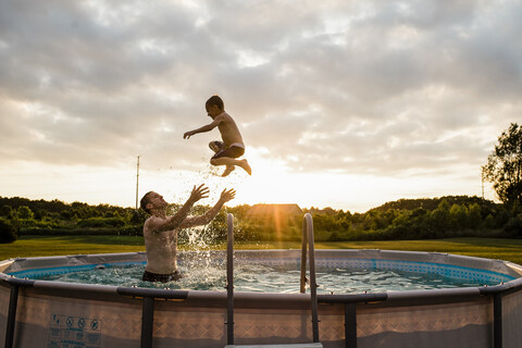 Vater fängt Sohn beim Schwimmen im Planschbecken gegen den bewölkten Himmel im Hinterhof bei Sonnenuntergang, lizenzfreies Stockfoto